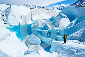 Young woman standing near deep blue lake on the Matanuska Glacier in Alaska. She wears a backpack and helmet with ice axe in hand