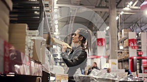 Young woman is standing near shelves with goods, looking for the necessary box, holding in her hands in a warehouse