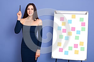 Young woman standing near sticky notes on flip chart, holding pen in raising hand, having great business idea, posing in dress