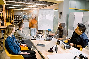 Young woman standing near the white office board with chart and speaking to her colleagues . Multiracial team of people