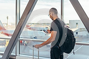 Young woman standing near window at airport holding mobile phone