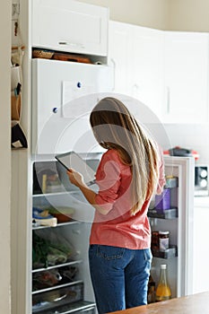 Young woman standing next to an opened refrigerator door, holding a smartphone and ordering fresh fruit and vegetables