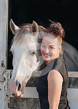 Young woman standing next to stable box with white Arabian horse inside
