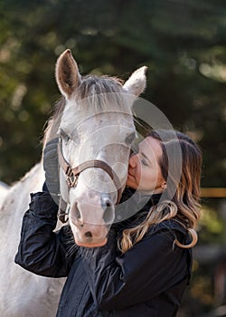 Young woman standing next to white Arabian horse eyes closed as if she\'s kissing or smelling, blurred trees background