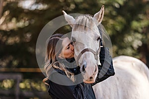 Young woman standing next to white Arabian horse eyes closed as if she's kissing or smelling, blurred trees background