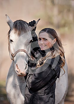 Young woman standing next to white Arabian horse, holding it by ears, sun shines on them, closeup detail