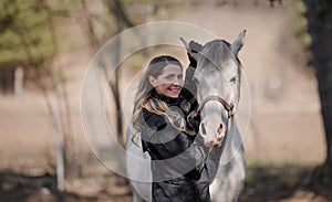 Young woman standing next to white Arabian horse, holding it by ears, sun shines on them, closeup detail