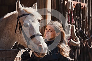 Young woman standing next to white Arabian horse resting in wooden stables box, sun shines on them, closeup detail
