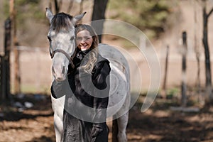 Young woman standing next to white Arabian horse smiling, sun shines on blurred trees and field background