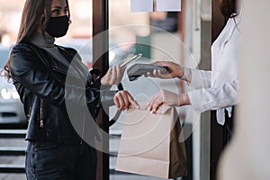 Young woman standing outdoors in mask and paying bill through smartphone using NFC technology in a cafe. Female customer