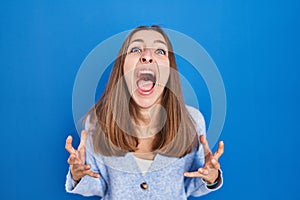 Young woman standing over blue background crazy and mad shouting and yelling with aggressive expression and arms raised