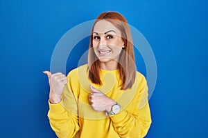 Young woman standing over blue background pointing to the back behind with hand and thumbs up, smiling confident