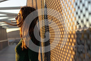 A young woman standing on a pedestrian bridge against steel mesh in sunset