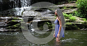 Young woman standing in the river near waterfall and looking at the falling water. Woman is smiling and breathing deeply