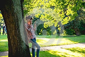 Young woman is standing under a tree with a phone in hands in th