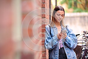 Young Woman Standing By Wall Looking At Mobile Phone Checking Text Messages And Social Media