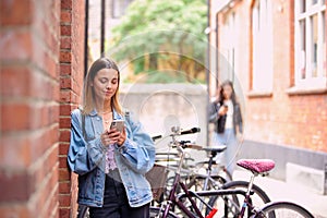 Young Woman Standing By Wall Looking At Mobile Phone Checking Text Messages And Social Media