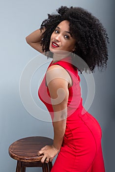 Young woman, standing, wearing a red outfit with curly hair posing next to a wooden stool