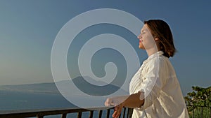 Young woman stands on a balcony with a beautiful view of the sea and mountains. Woman gladly exposes her face to the
