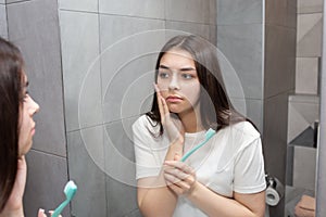 A young woman stands in the bathroom and holds her cheek because of a toothache. Girl frowns in pain. Importance of oral