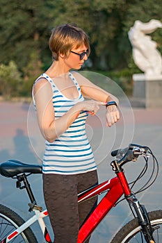 Young woman stands with a bicycle and looks at her fitness brace