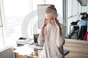 Young woman stands in a big office room and keeping her hands close to the head. She has a headache. It is a strong pain