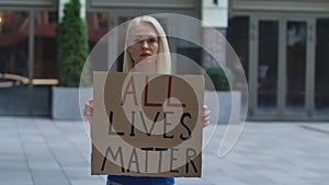 Young woman stands with a cardboard poster ALL LIVES MATTER in a public place outdoor. A protesting political activist