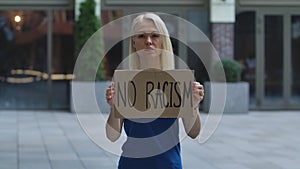 Young woman stands with a cardboard poster NO RACISM in a public place outdoor. A protesting political activist at a