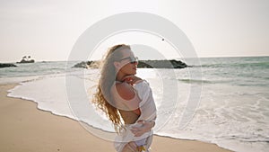Young woman stands, curly long hair blowing in wind at sandy beach, sea surf. Attractive model in sunglasses posing