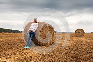 A young woman stands on the field, leaning on a sheaf of hay.