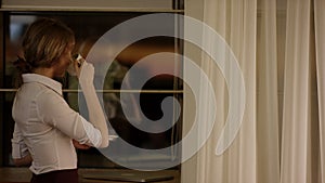 Young woman stands in front of the window and drinks a cup of tea or coffee