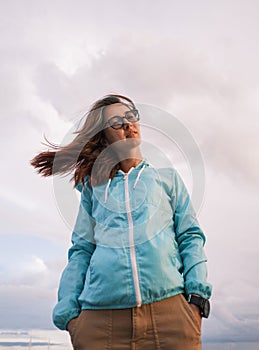 A young woman stands in glasses and a blue windbreaker, her hair fluttering in the wind.