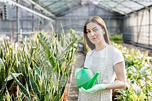 A young woman stands in the middle of a large greenhouse holding a watering can and smiling at the camera. The concept of caring