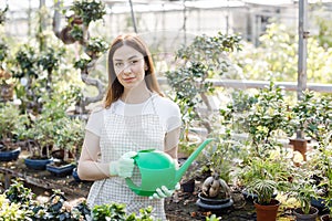 A young woman stands in the middle of a large greenhouse holding a watering can and smiling at the camera. The concept of caring