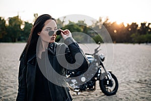 Young woman stands near black motorbike on beach at sunset