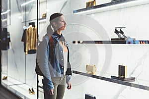 Young woman stands near store window of the boutique, looks at handbags and shoes