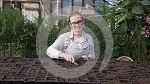 Young woman stands near table with seedlings in large greenhouse and holds rake and shovel in her hands. Gardening