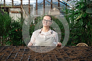 Young woman stands near table with seedlings in large greenhouse and holds rake and shovel in her hands. Gardening