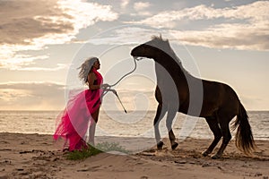 Young woman stands in a pink dress on a beach, with a horse beside her