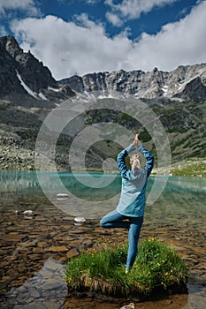 Young woman stays in one leg yoga position against the turquoise lake in the mountains