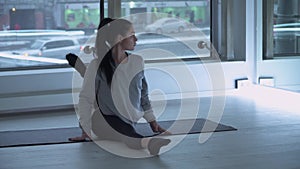 Young woman stratching legs on mat in the gym with large floor-to-ceiling windows. Modern urban cityscape, cars behind