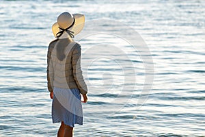 Young woman in straw hat and a dress standing alone on empty sand beach at sea shore. Lonely tourist girl looking at horizon over