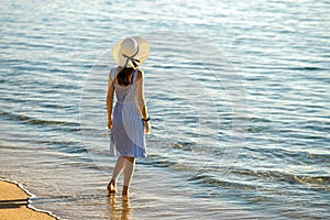 Young woman in straw hat and a dress standing alone on empty sand beach at sea shore. Lonely tourist girl looking at horizon over