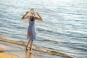 Young woman in straw hat and a dress standing alone on empty sand beach at sea shore. Lonely tourist girl looking at horizon over