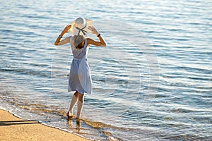 Young woman in straw hat and a dress standing alone on empty sand beach at sea shore. Lonely tourist girl looking at horizon over