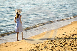 Young woman in straw hat and a dress walking alone on empty sand beach at sea shore. Lonely tourist girl looking at horizon over