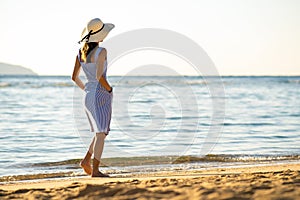 Young woman in straw hat and a dress walking alone on empty sand beach at sea shore. Lonely tourist girl looking at horizon over