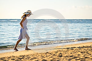 Young woman in straw hat and a dress walking alone on empty sand beach at sea shore. Lonely tourist girl looking at horizon over