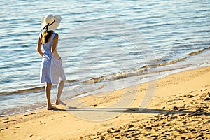 Young woman in straw hat and a dress walking alone on empty sand beach at sea shore. Lonely tourist girl looking at horizon over