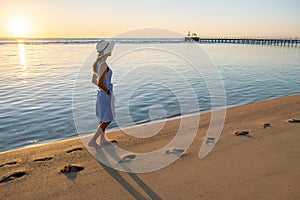 Young woman in straw hat and a dress walking alone on empty sand beach at sunset sea shore. Lonely girl looking at horizon over